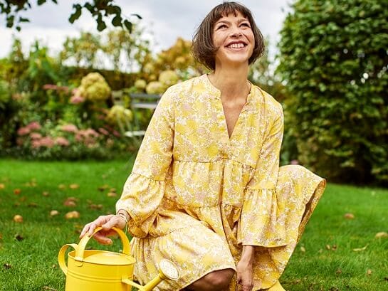 Eine Frau in einem gelben Sommerkleid sitzt fröhlich auf dem Rasen in ihrem Garten und hält in ihrer rechten Hand eine gelbe Gießkanne
